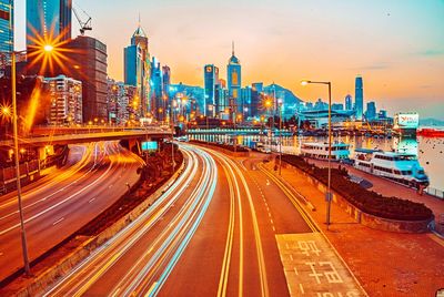 Light trails on road by buildings against sky at dusk