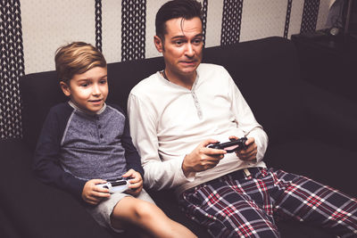 Father and son playing video games while sitting on sofa