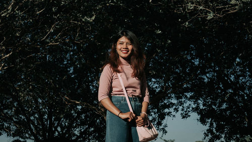 Smiling young woman carrying purse while standing against trees