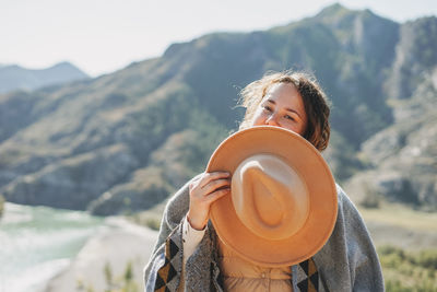 Carefree brunette young woman traveler in poncho with felt hat on background of mountain river