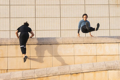 Full body male athletes in sportswear and sneakers parkour jumping above stone roof border while exercising on sunny day on street