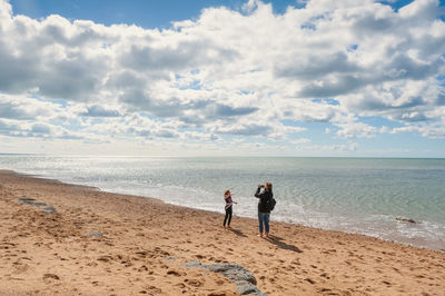 Full length of mother photographing daughter while standing on beach against sky