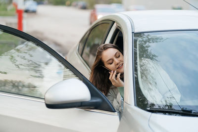 Beautiful girl with long hair in a grey trench coat using smartphone call gets into the car