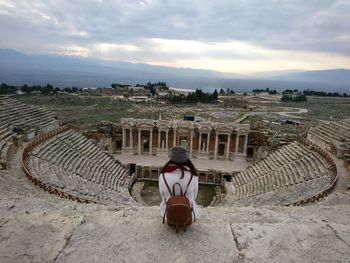 Rear view of woman sitting at amphitheater against cloudy sky during sunset