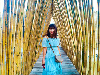 Portrait of smiling young woman standing on boardwalk amidst bamboos