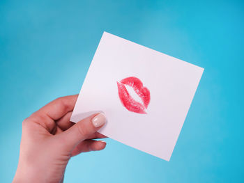 Cropped hand of woman holding paper with lipstick kiss against blue background