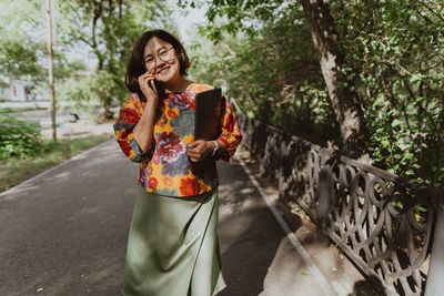 Happy woman chatting on the phone holding a laptop walking in the park