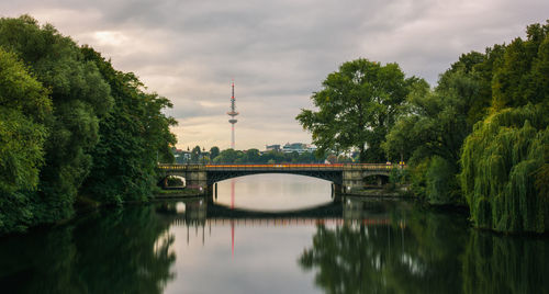 Bridge over river amidst trees against cloudy sky