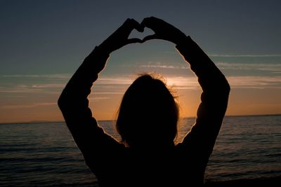 Silhouette woman making heart shape against sea during sunset