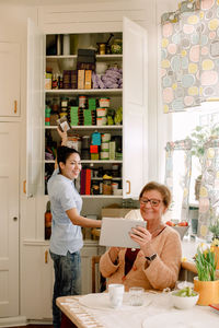 Smiling elderly woman showing digital tablet to young female caregiver working in kitchen at nursing home