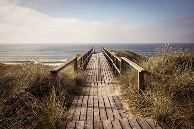 Empty boardwalk leading to calm sea against cloudy sky