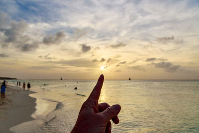 Cropped hand of person pointing at sun on shore against cloudy sky during sunset