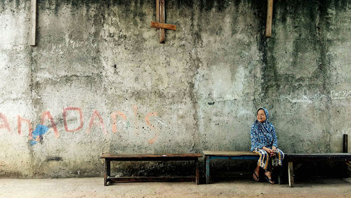 Full length of senior woman sitting on bench against wall