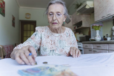 Senior woman with eyeglasses counting money on documents at home