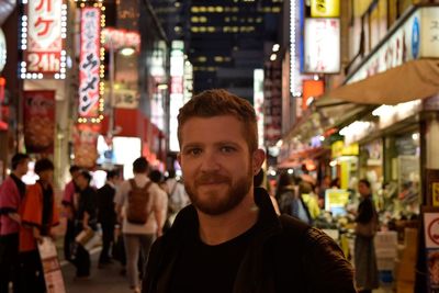 Portrait of smiling man standing amidst illuminated market in city at night