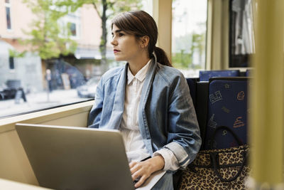 Young woman looking through tram window while using laptop