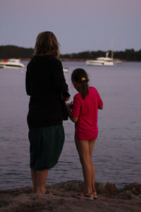 Rear view of mother and daughter standing on beach against clear sky
