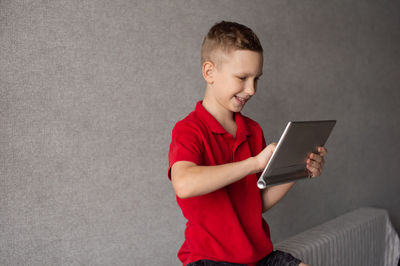 A cute boy in a red t-shirt looks at the tablet and laughs