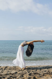 Side view of ethnic female leaning back while standing in ashta chandrasana pose during yoga practice on sandy coast against ocean