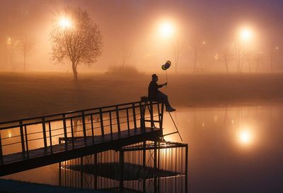 Man with balloons sitting on pier by calm lake in illuminated park at night