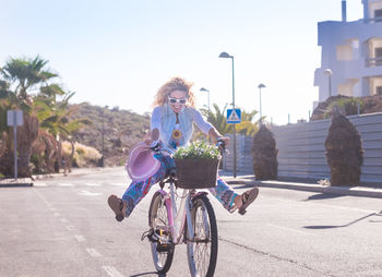 Full length of cheerful woman riding bicycle on road against sky in city