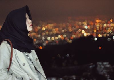 Side view of woman wearing hijab standing against illuminated lights at night