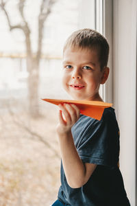 A boy with an orange paper airplane, sits at the window and looks out into the street. 