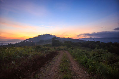 Dirt road amidst field against sky during sunset