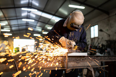 Unrecognizable male employee in protective gloves and helmet using welding machine while working in dark workshop