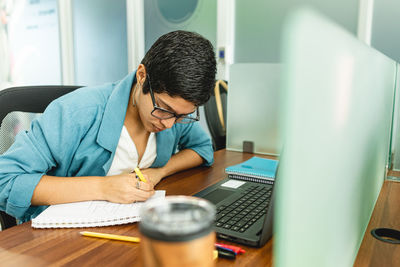 Hispanic woman with short hair in eyeglasses and stylish clothes sitting at wooden table with laptop and writing notes in notebook while working on project in office in costa rica