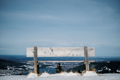 Wooden bench on snow covered land against sky