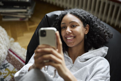 Smiling young woman lying on bean bag and using phone