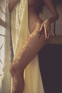 Midsection of naked young woman standing by window curtain at home