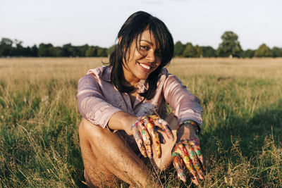 Smiling woman with paint on hands sitting at park during sunny day