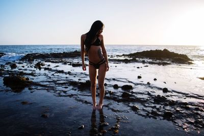 Full length of young woman in bikini standing at beach against clear sky