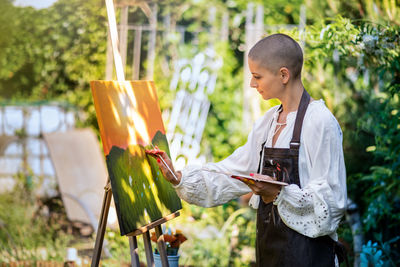 Beautiful young woman relaxing while painting an art canvas outdoors in her garden. 