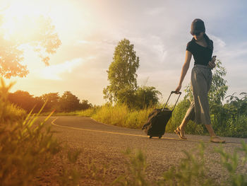 Woman pulling suitcase on road against sky during sunset