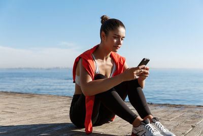 Young woman using phone while sitting on beach against sky