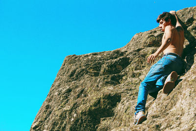 Low angle view of shirtless young man hanging on cliff