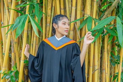 Woman wearing graduation gown standing against bamboos