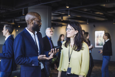 Side view of smiling male with smart phone talking to female colleague in office