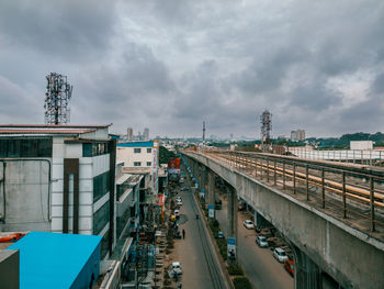 Railway bridge by buildings in city against cloudy sky