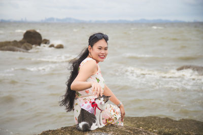 Smiling woman with cat sitting on rock at beach