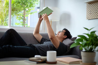 Young man reading book while lying on sofa at home