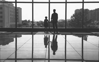 Rear view of father and daughter against glass in building with reflection on floor