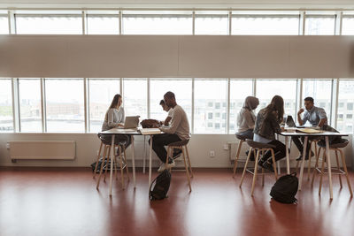 Full length of multi-ethnic students studying at desks by window in high school cafeteria