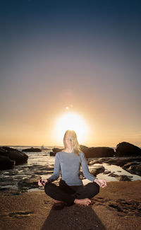 Back lit woman meditating yoga on shore at beach against sky during sunrise