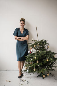 Happy woman standing with arms crossed by damaged christmas tree in front of white wall