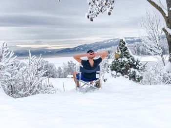 Rear view of man relaxing on snowcapped mountain against cloudy sky