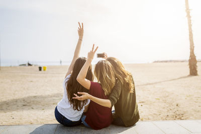 Rear view of three female friends sitting at the beach taking a selfie